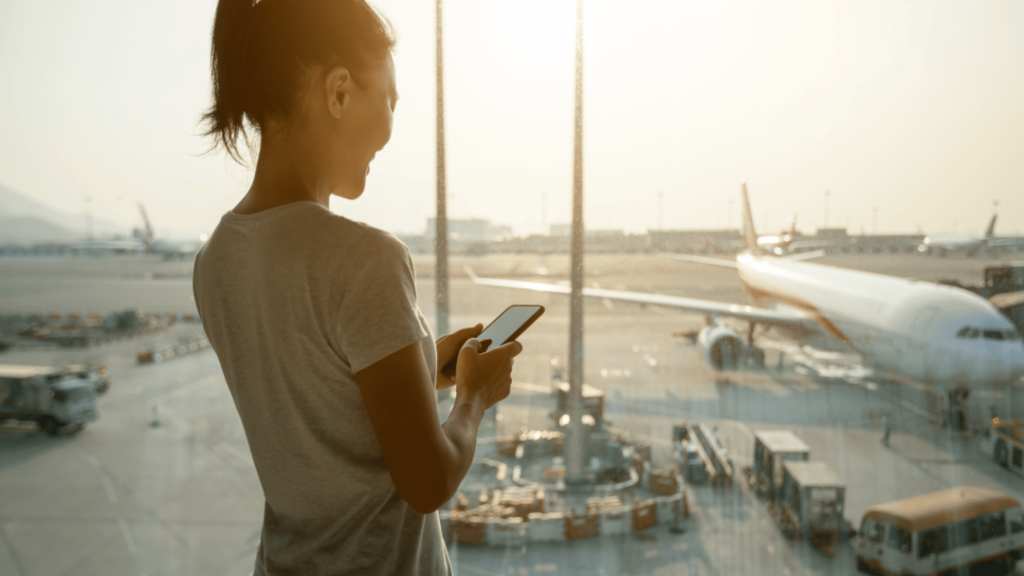 A woman holding her phone looking out over an airport runway.
