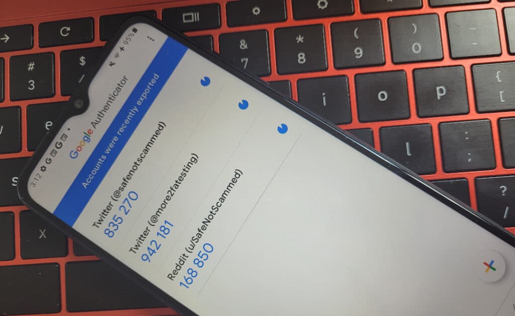 Google Authenticator on an Android phone displaying three 2FA codes for two Twitter accounts and a Reddit account. The phone is resting on a red laptop.