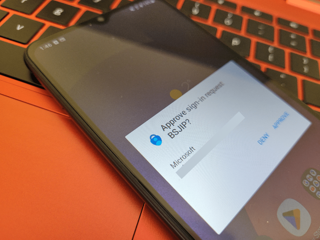A phone on a laptop with a push notification from Microsoft. It reads "Approve sign-in request BSJIP?" It lists the Microsoft account email, which is redacted, and gives two options: Deny and Approve.