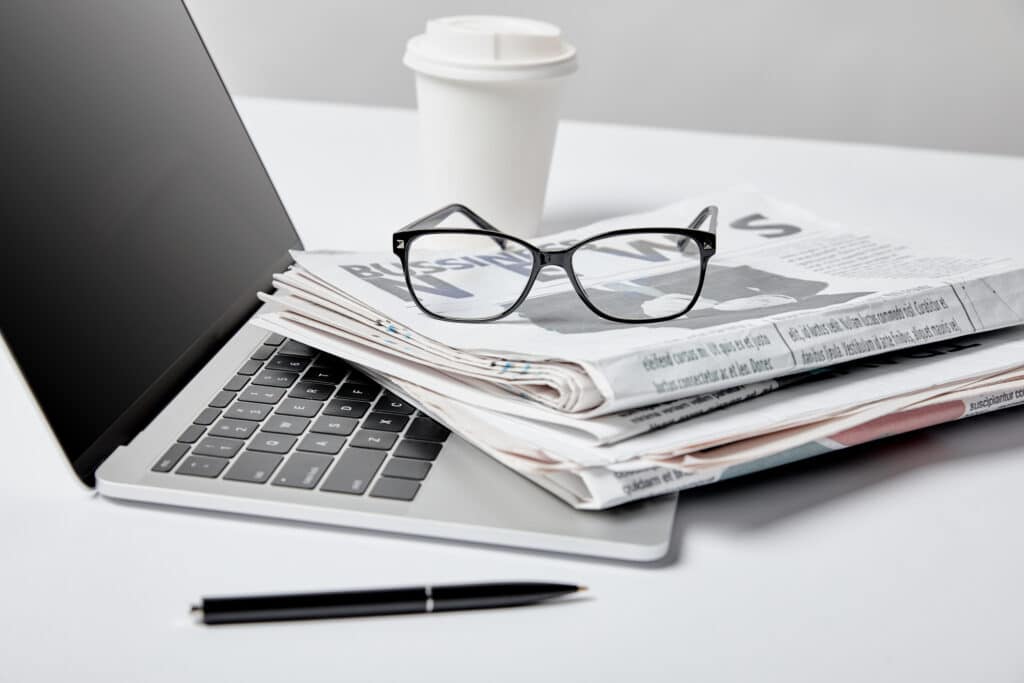 An open laptop with multiple newspapers on its keyboard and a pair of glasses. A coffee cup is in the background.