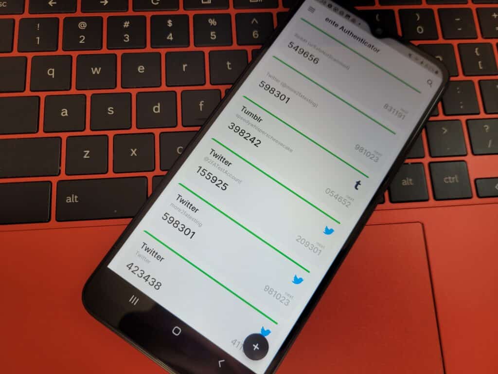 A photo of ente Auth, installed on my backup phone and sitting on my red Chromebook. It is displaying codes for six different accounts.