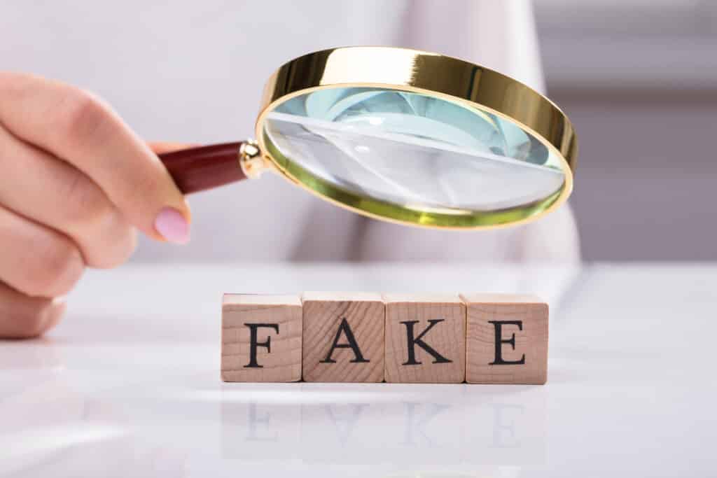 A person holds a magnifying glass and is using it to examine wooden blocks that spell out "fake."