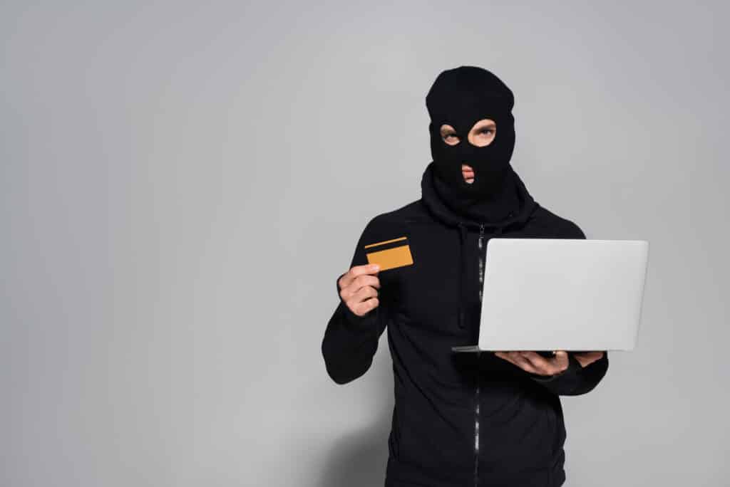 A man in a black balaclava holding a laptop and a credit card.