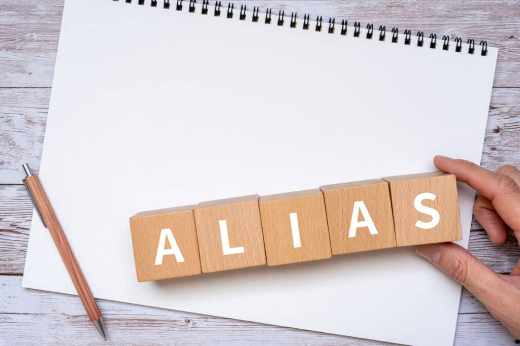 The word 'ALIAS" is spelled out using wooden blocks on top of a piece of paper. A pen lies to the left of the blocks.